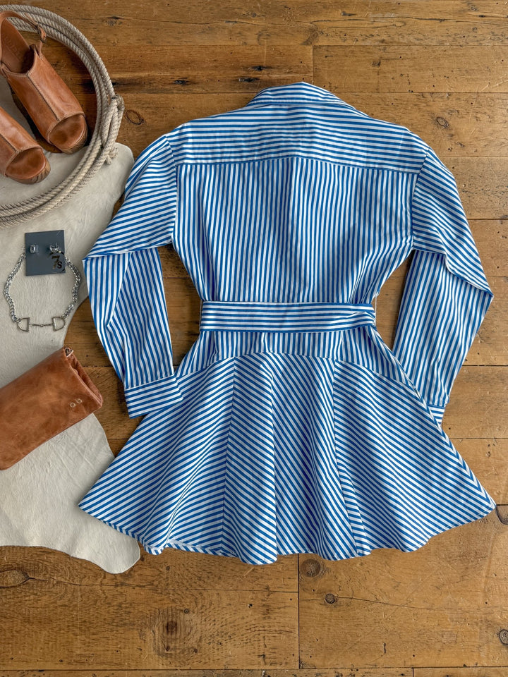 The Laneville Blue Striped Button Up Dress by Savannah Sevens western life{&}style is laid out on a wooden floor, surrounded by brown sandals, a tan suede clutch, a silver bracelet, and a white cloth.