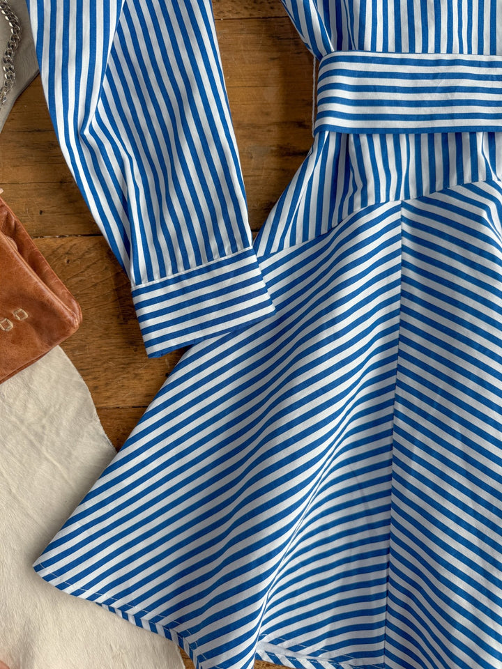 The Laneville Blue Striped Button Up Dress by Savannah Sevens western life{&}style, featuring blue and white stripes, long sleeves, and a belt, is laid out on a wooden surface beside a brown bag and a piece of white fabric.