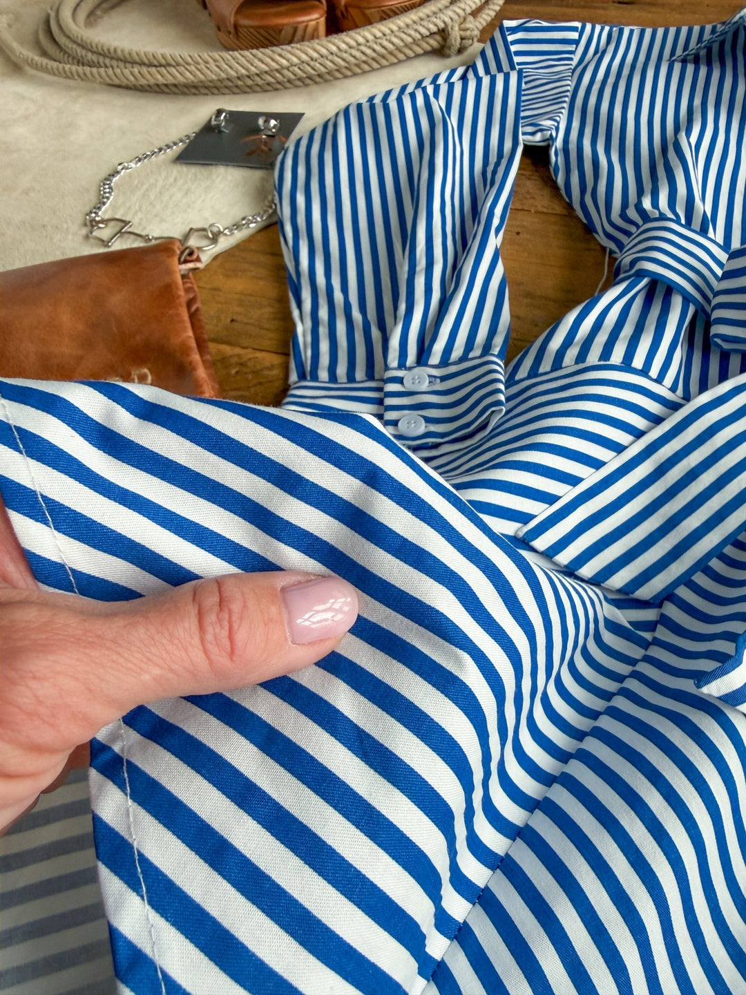 A hand with pale pink nails holds the sleeve of the Laneville Blue Striped Button Up Dress by Savannah Sevens western life{&}style, with a tan leather bag, chain, and rope in the background on a wooden and beige surface.