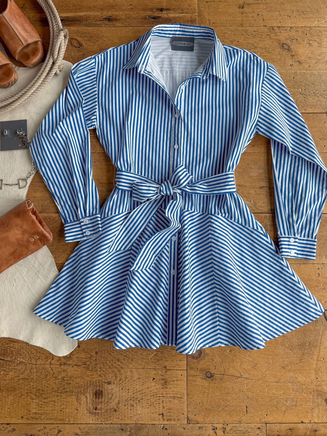The Laneville Blue Striped Button Up Dress by Savannah Sevens western life{&}style is a blue and white, vertically striped, collared cotton dress with long sleeves and a waist tie, shown laid flat on a wooden floor. Styled alongside sandals, boots, a rope, and a cowhide rug for a Western-inspired look.