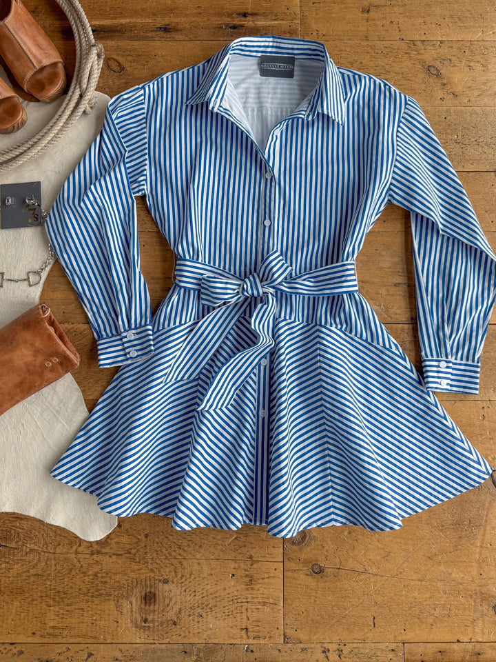 The Laneville Blue Striped Button Up Dress by Savannah Sevens western life{&}style is a blue and white, vertically striped, collared cotton dress with long sleeves and a waist tie, shown laid flat on a wooden floor. Styled alongside sandals, boots, a rope, and a cowhide rug for a Western-inspired look.