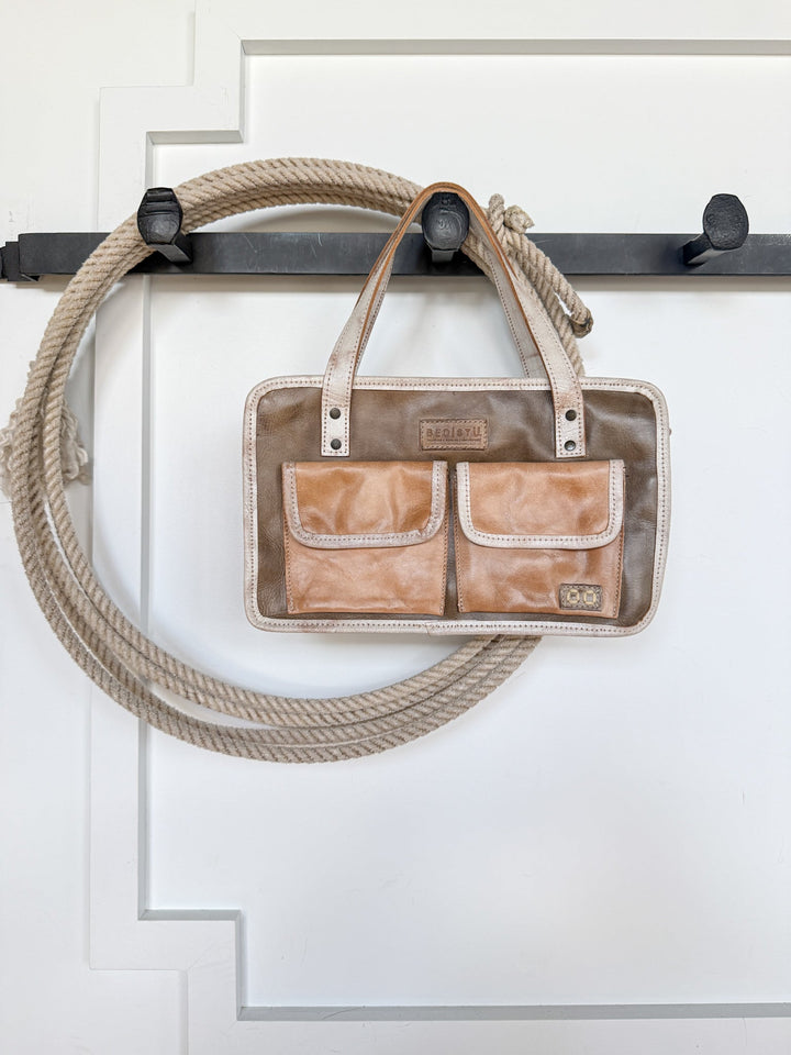 The Lanexa Stone Leather Handbag by Savannah Sevens western life{&}style, featuring a tan and brown genuine leather design with two front pockets, hangs on a black hook alongside a coiled rope against a white paneled wall.