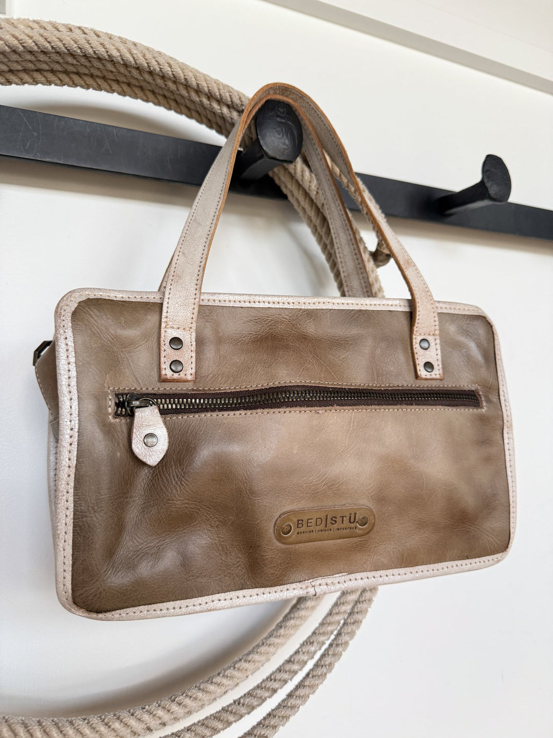 A tan Lanexa Stone Leather Handbag by Savannah Sevens western life{&}style, featuring a front zipper pocket, hangs on a black wall hook with coiled rope in the background against a white wall.