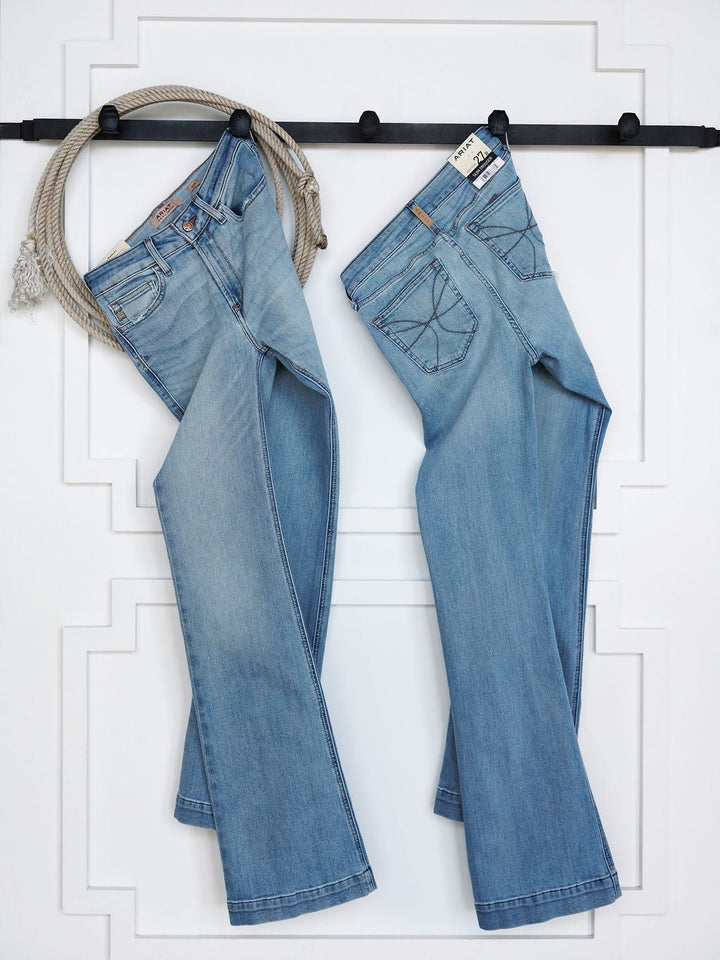 Two pairs of Savannah Sevens western life{&}style The Langdon High Rise Slim Trouser Jeans in medium light wash hang from black hooks on a white wall, with a rope beside one pair; front and back views are shown.