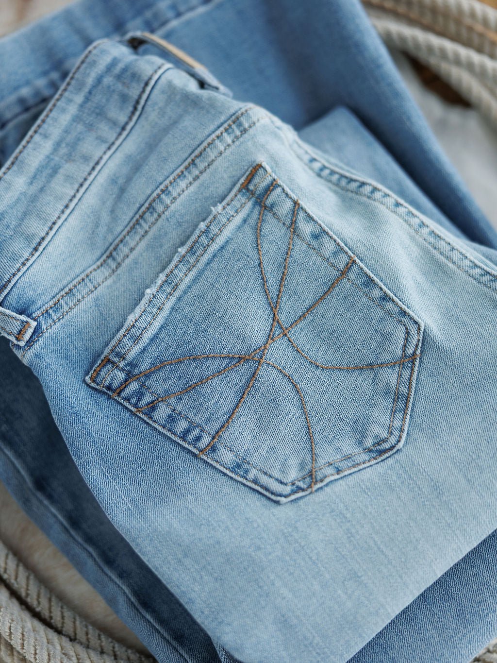 A close-up of Savannah Sevens western life{&}style's The Langdon High Rise Slim Trouser Jean in medium light wash denim shows the back pocket’s brown stitching, with another pair underneath highlighting the high-rise texture and details.