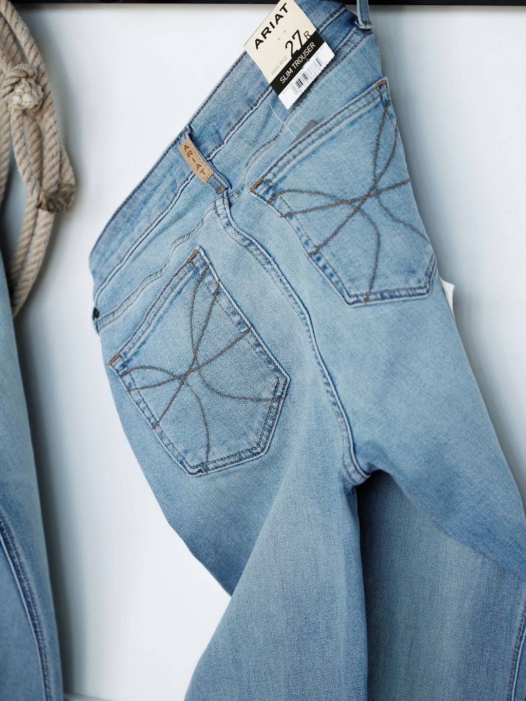 Light blue medium wash The Langdon High Rise Slim Trouser Jeans by Savannah Sevens western life{&}style hang, showing star-stitched back pockets. A tag is visible, and a coiled rope appears partially to the left.