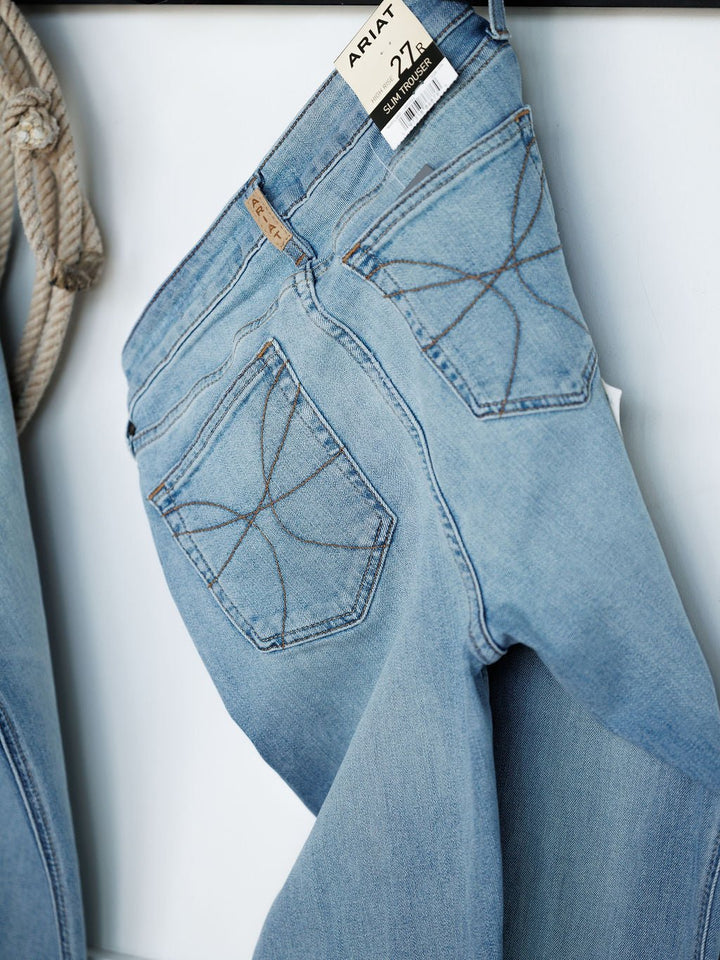 Light blue medium wash The Langdon High Rise Slim Trouser Jeans by Savannah Sevens western life{&}style hang, showing star-stitched back pockets. A tag is visible, and a coiled rope appears partially to the left.