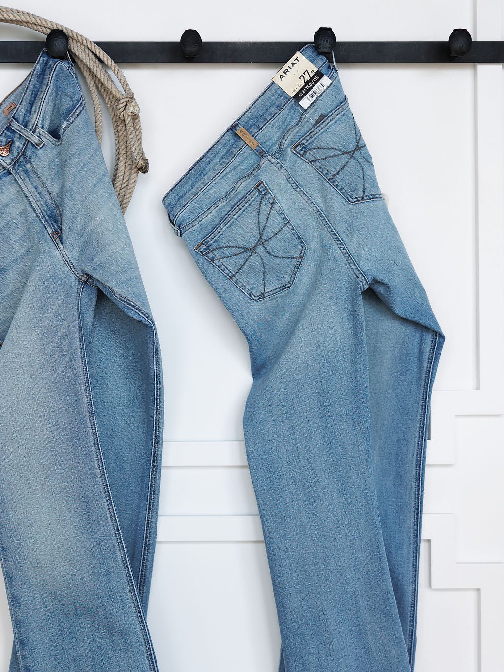 Two pairs of Savannah Sevens western life{&}style The Langdon High Rise Slim Trouser Jean in medium light wash hang on black hooks against a white wall, with a coiled rope beside them; one pair shows the back pocket and waistband tag.