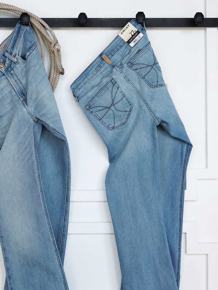Two pairs of Savannah Sevens western life{&}style The Langdon High Rise Slim Trouser Jean in medium light wash hang on black hooks against a white wall, with a coiled rope beside them; one pair shows the back pocket and waistband tag.