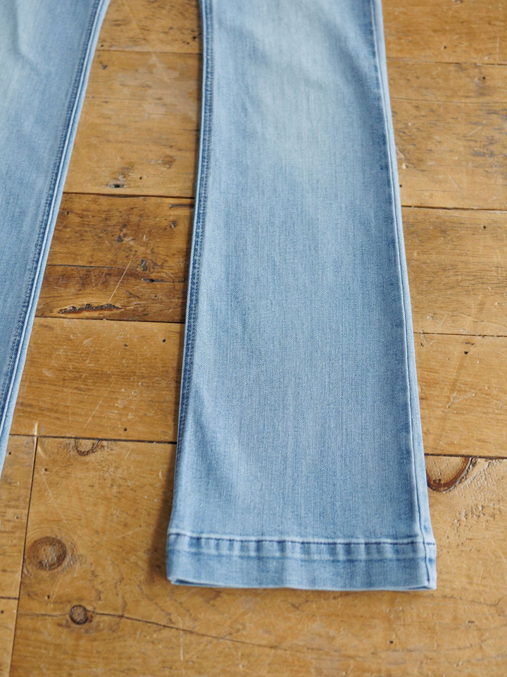 A close-up of The Langdon High Rise Slim Trouser Jean by Savannah Sevens western life{&}style in medium light wash denim, showcasing straight leg cut and hem details, laid flat on a wooden floor.