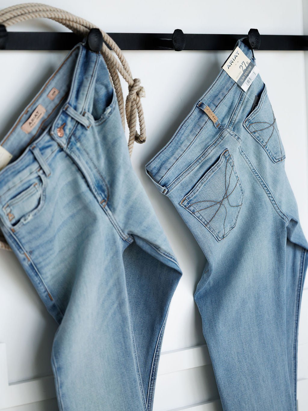 Two pairs of The Langdon High Rise Slim Trouser Jeans by Savannah Sevens western life{&}style hang on black hooks against a white wall, one with a rope draped over the waistband and the other showing a size tag on the belt loop.