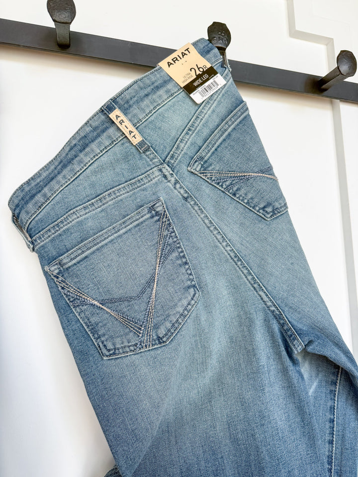 A pair of Savannah Sevens western life{&}style Langston High Rise Wide Leg Jeans in a medium light wash hangs on a black hook. These ultra high rise wide leg jeans feature two back pockets with stitching details and a visible tag showing the size, 26.