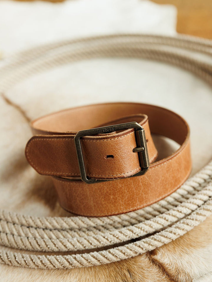 The Langston Belt by Savannah Sevens western life{&}style, crafted from genuine light brown leather with a metal buckle, is rolled up atop a coiled rope on a hide surface.