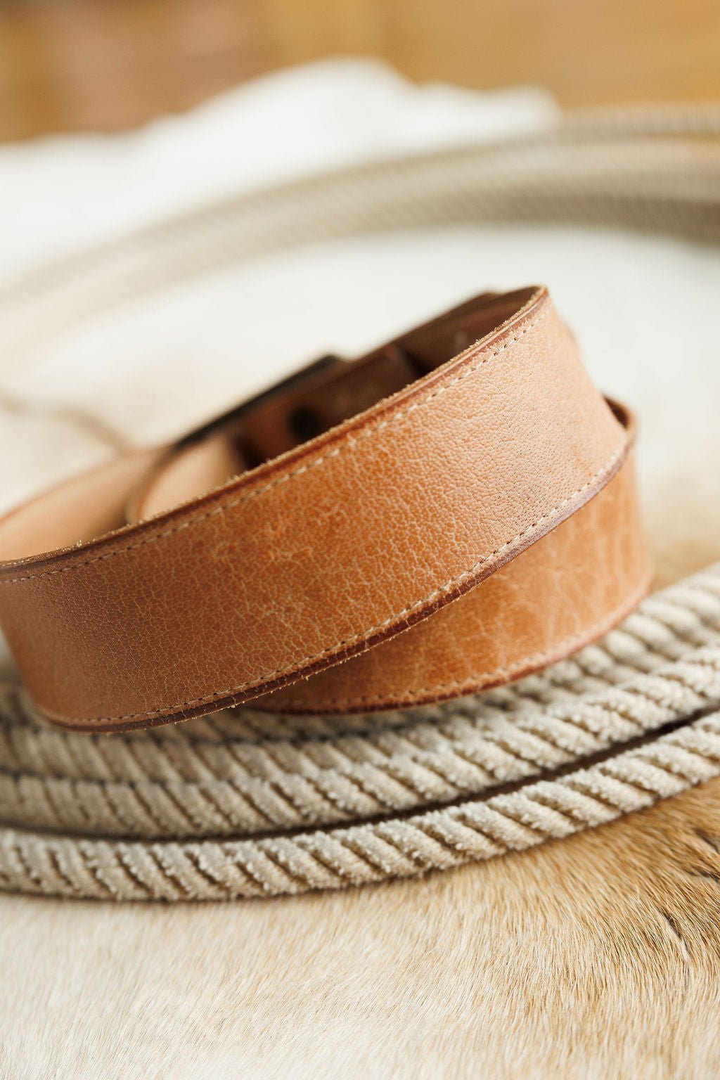 A close-up of the Langston Belt by Savannah Sevens western life{&}style, crafted from brown genuine leather with visible texture, natural creases, and an interchangeable buckle, resting on a coiled rope atop a light surface.