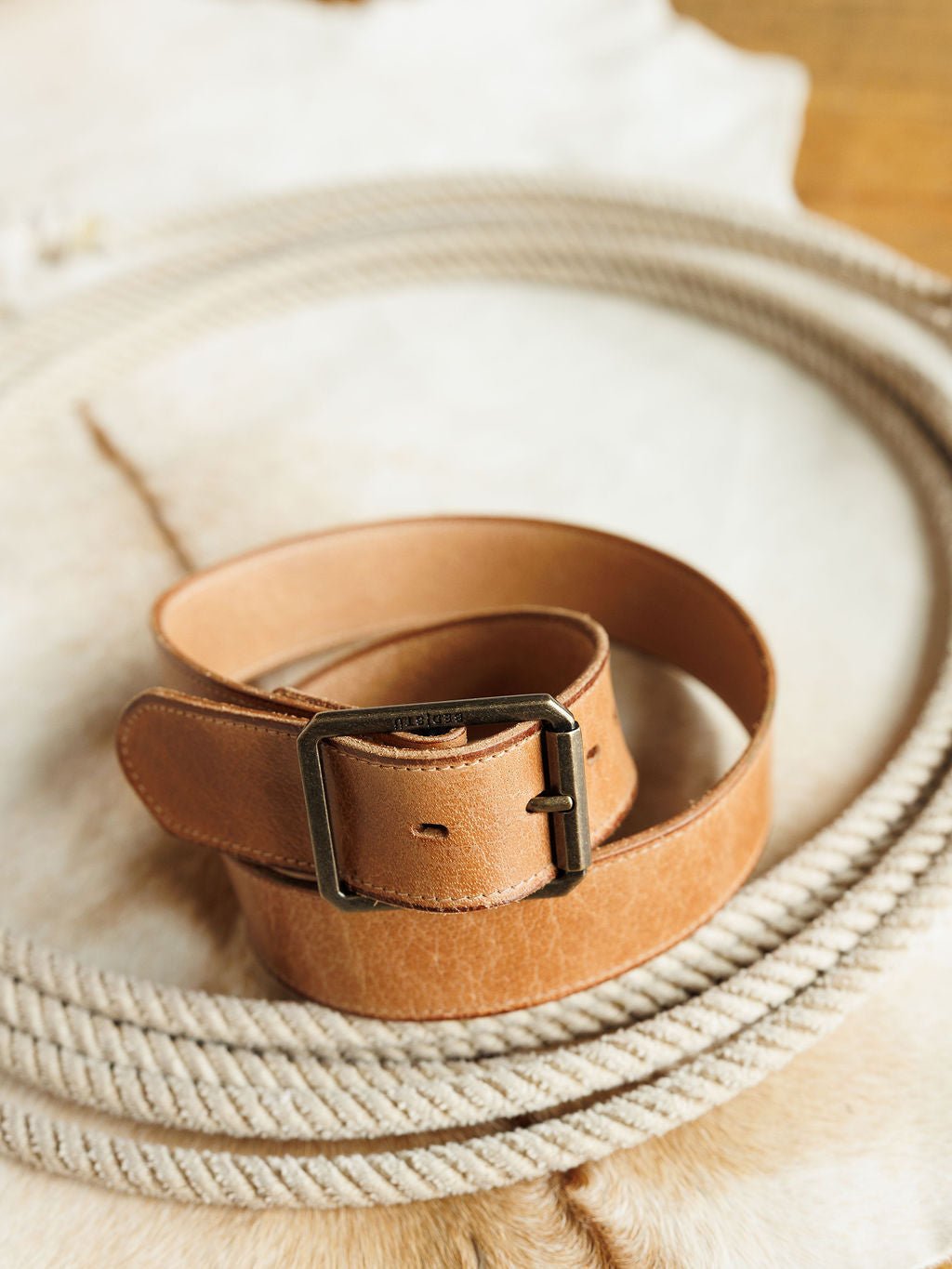 The Langston Belt by Savannah Sevens western life{&}style, crafted from genuine tan leather and featuring a rectangular metal buckle, is displayed coiled on rope against a light cowhide backdrop, blending style with durability.