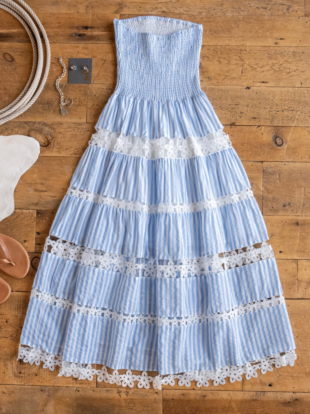 The Lantana Blue Striped Strapless Dress by Savannah Sevens western life{&}style is laid flat on a wooden floor, accented with jewelry, tan sandals, and a soft white rug.
