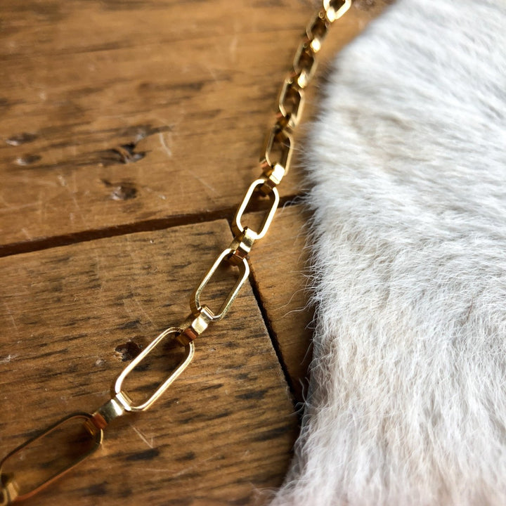 The Larkin Bracelet by Savannah Sevens western life{&}style, an 18K gold filled piece with rectangular links, is displayed on wood next to light-colored cowhide.
