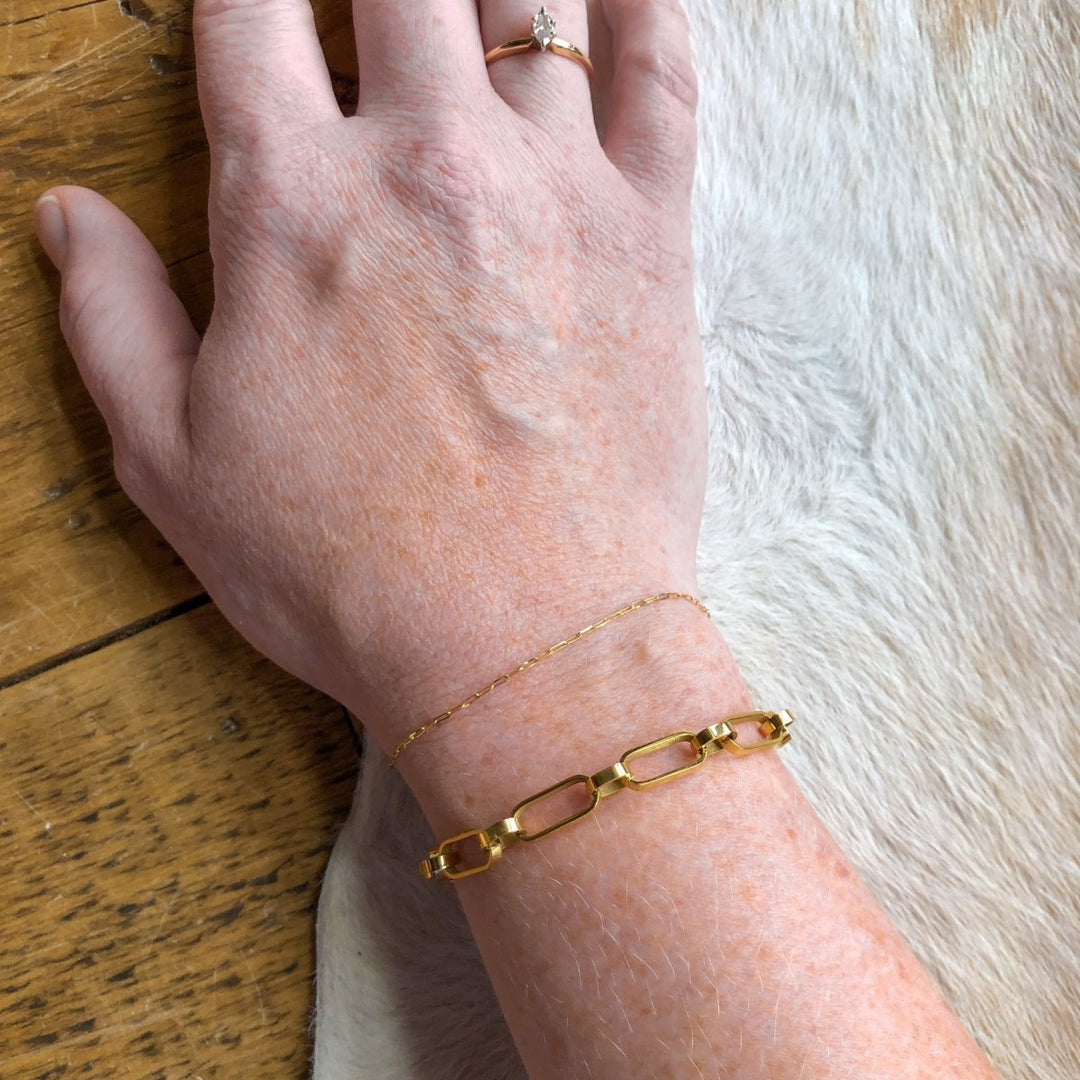 A hand wearing an 18K gold-filled ring with a clear stone and two bracelets—including the bold Larkin Bracelet by Savannah Sevens western life{&}style—rests on light fur beside a wooden table. All pieces are hypoallergenic.