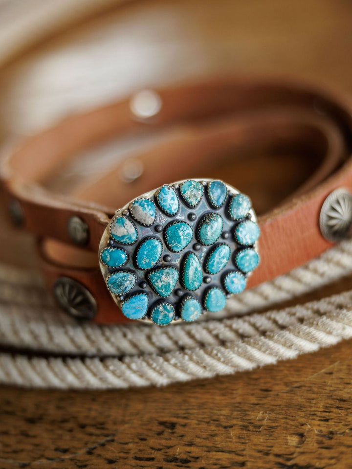 The Las Cruces Buckle by Savannah Sevens western life{&}style is a brown leather belt featuring a large oval buckle adorned with multiple Number 8 Turquoise stones, shown resting on a coiled white rope atop a wooden surface.