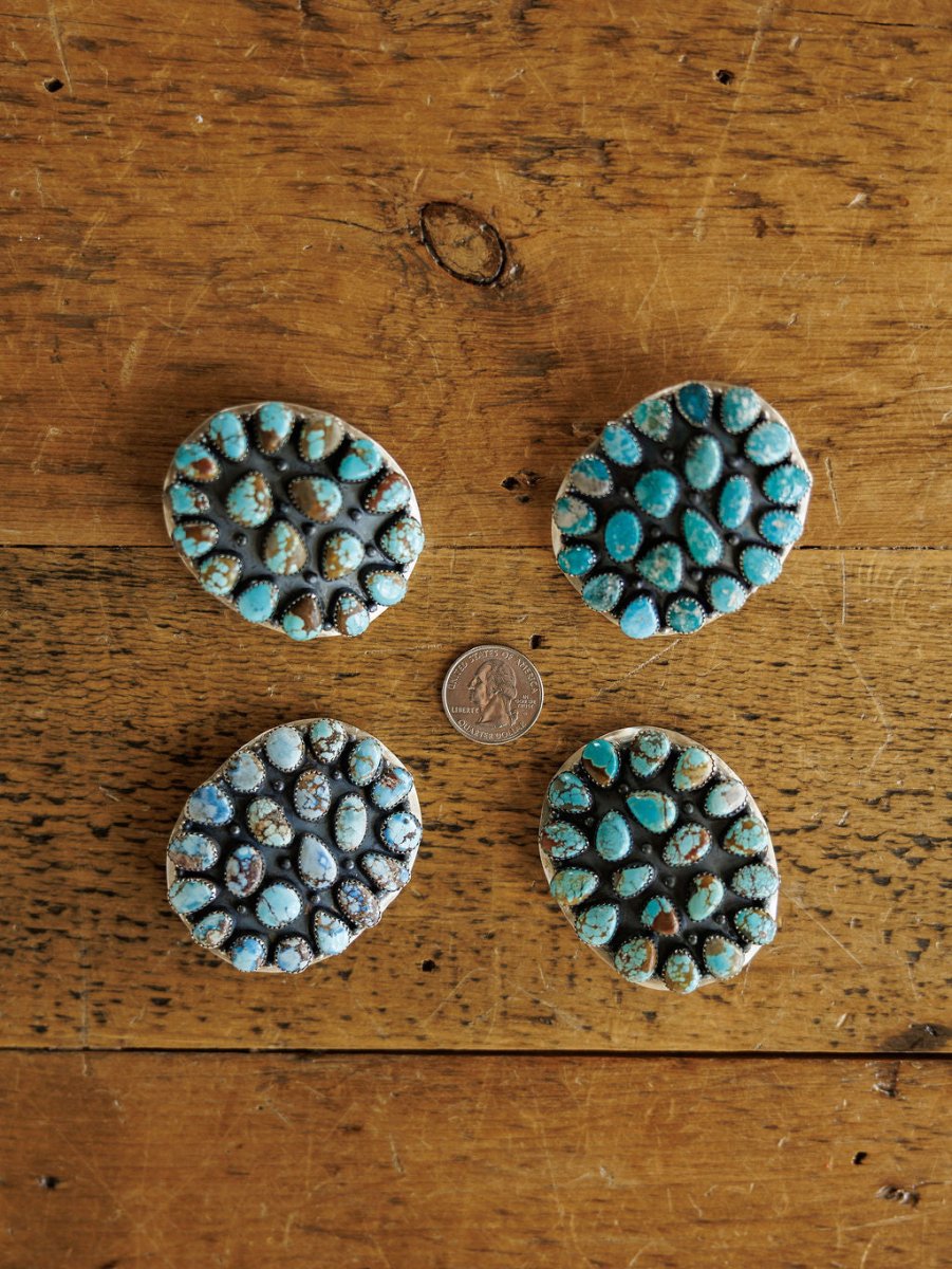 Four Las Cruces Buckles by Savannah Sevens western life{&}style, each with clustered Number 8 Turquoise stones, form a circle around a US quarter on a rustic wooden surface.