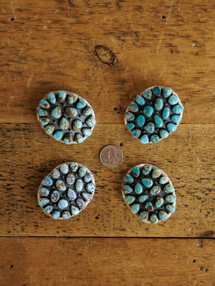 Four Las Cruces Buckles by Savannah Sevens western life{&}style, each with clustered Number 8 Turquoise stones, form a circle around a US quarter on a rustic wooden surface.