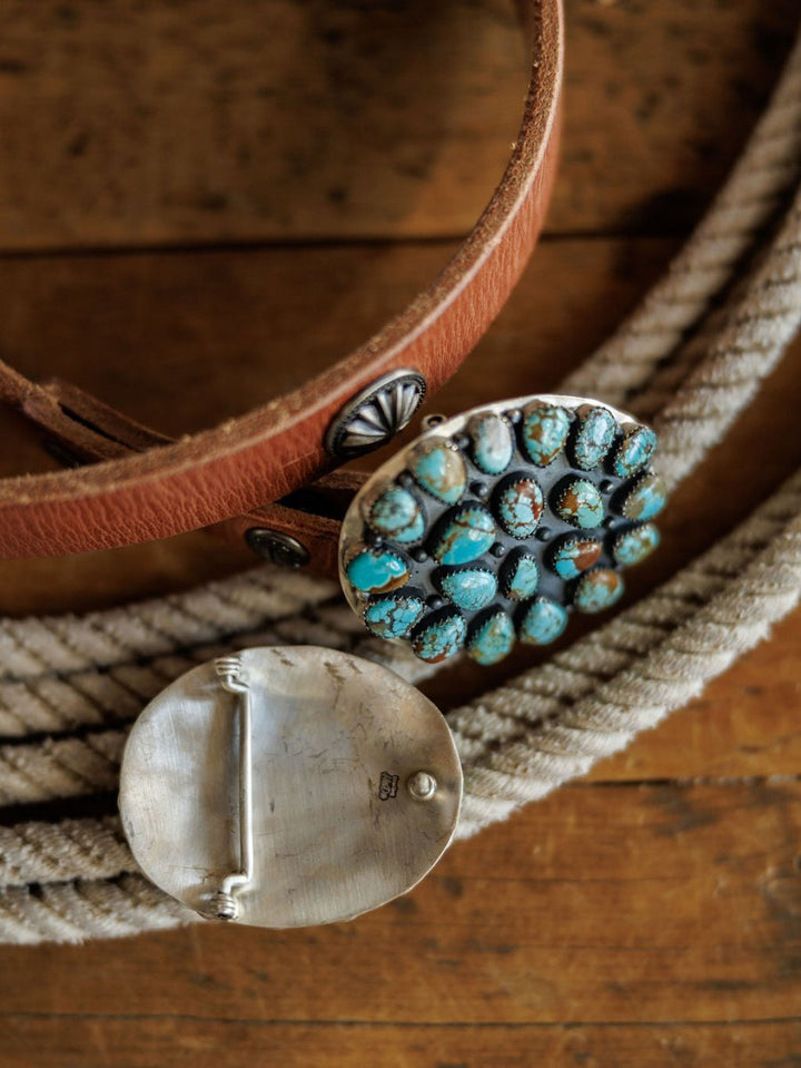 A close-up of the Savannah Sevens western life{&}style Las Cruces Buckle, a large oval sterling silver buckle with Number 8 Turquoise on a brown leather belt, lying on coiled rope atop wood. The back of the buckle is also visible.