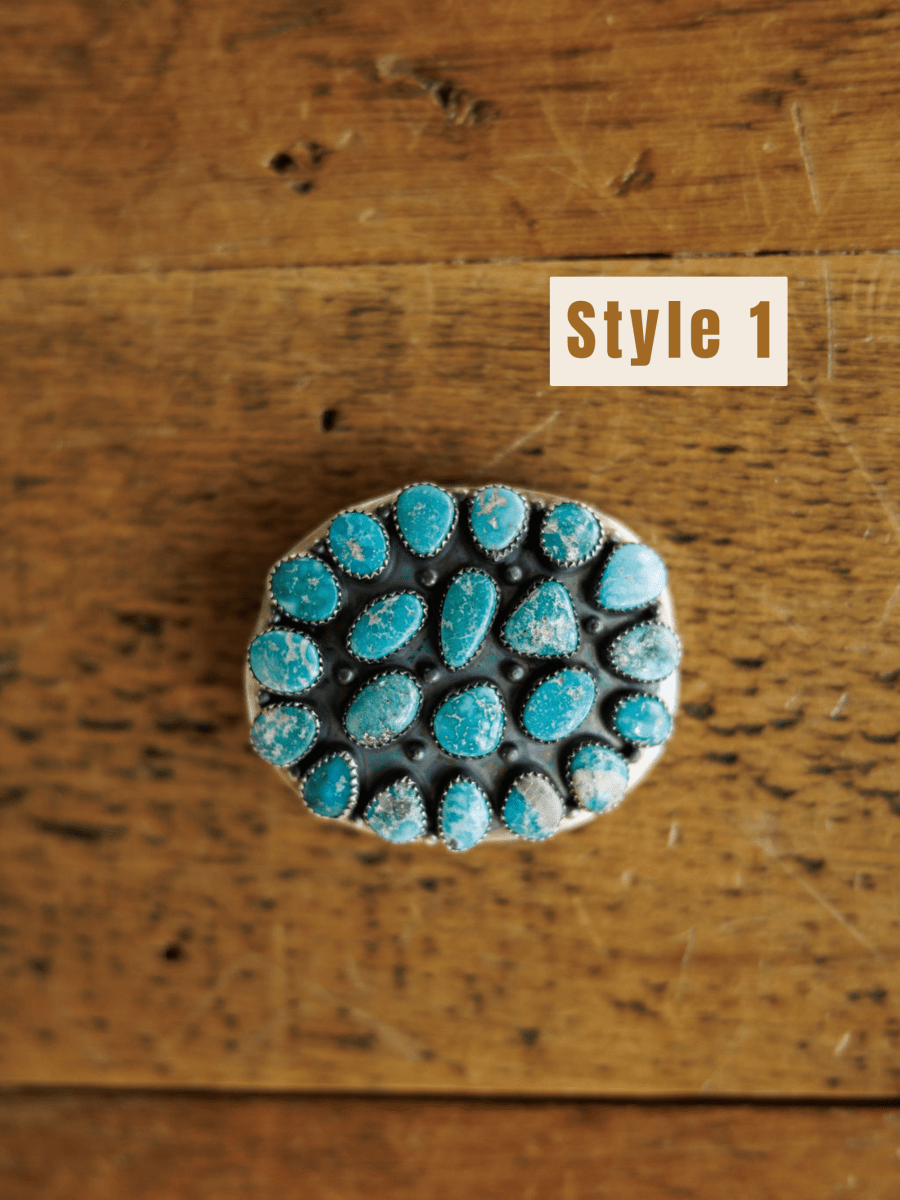 The Las Cruces Buckle by Savannah Sevens western life{&}style is shown on a rustic wooden surface, with "Style 1" labeled in the top right corner. Multiple oval White Water Turquoise stones accent its round silver design.