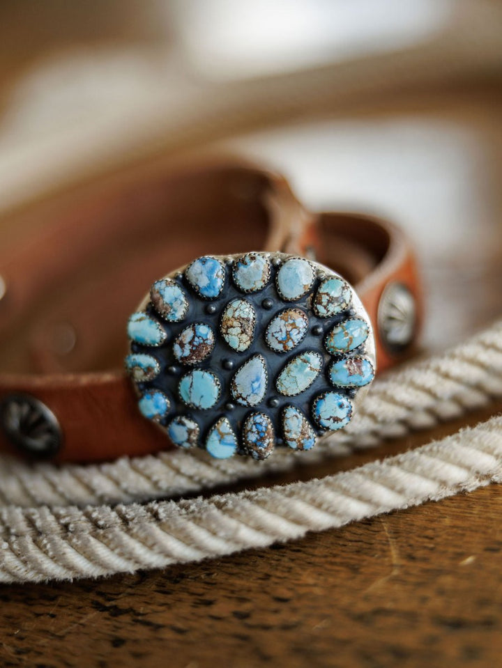 The Las Cruces Buckle by Savannah Sevens western life{&}style features multiple Number 8 Turquoise stones set in sterling silver on a brown leather belt, displayed on textured white rope over a wooden surface.