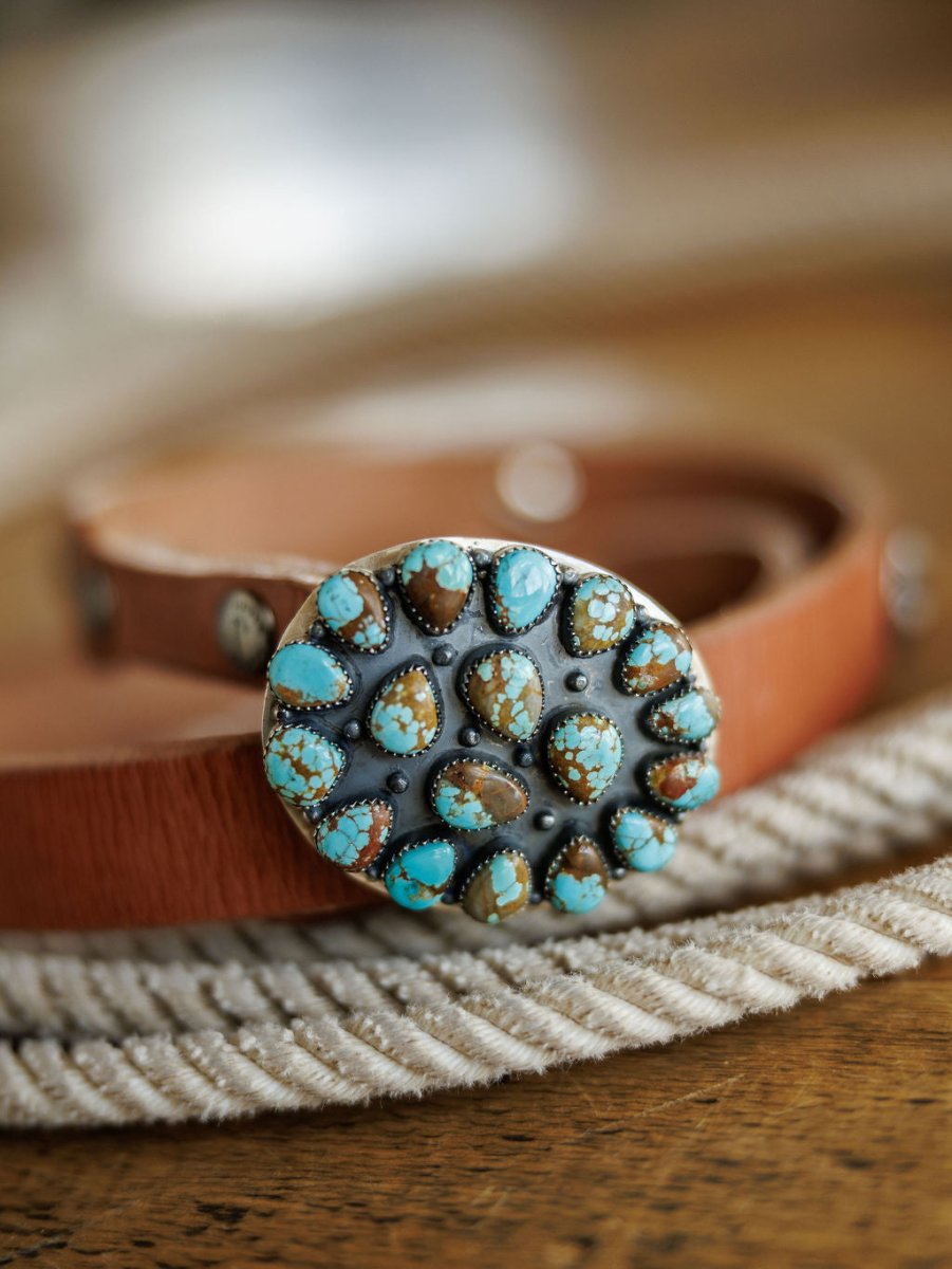 A close-up of the Las Cruces Buckle by Savannah Sevens western life{&}style, a large oval sterling silver buckle with Number 8 Turquoise and brown stones, shown on a brown leather belt resting atop rope on wood.