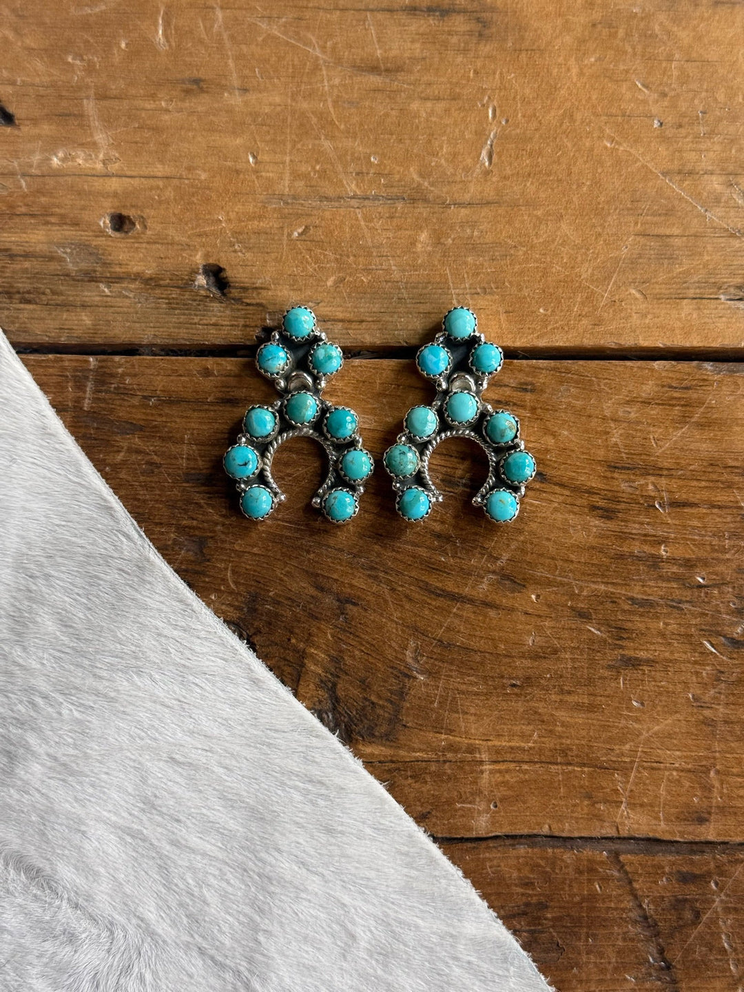 A pair of Savannah Sevens western life{&}style Latexo Turquoise Squash Earrings, crafted from sterling silver and featuring Kingman turquoise stones arranged in arches, displayed on a wooden surface beside a white textured fabric.