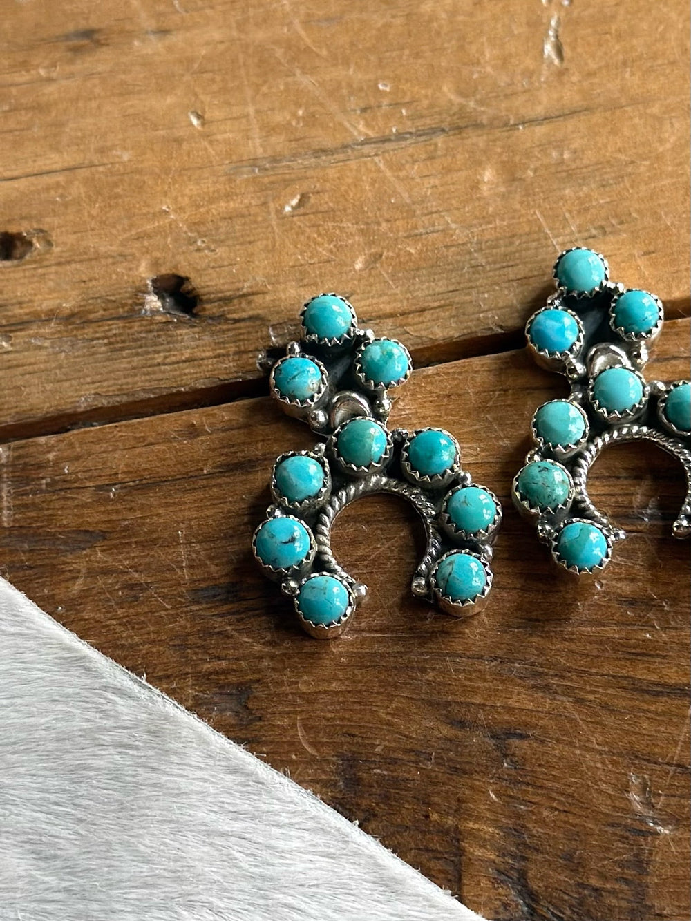 A pair of Savannah Sevens western life{&}style Latexo Turquoise Squash Earrings, crafted from sterling silver and adorned with multiple round Kingman turquoise stones arranged in a horseshoe shape, rests on a wooden surface with a white textured area in the corner.