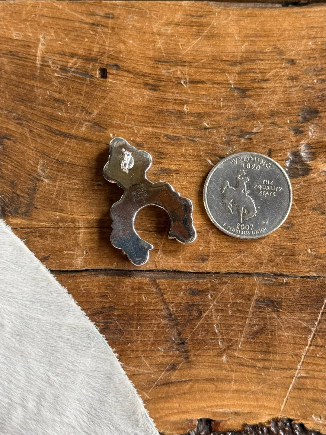 A small, squash with an open center lies next to a Wyoming state quarter on a wooden surface. A piece of light-colored cowhide is visible in the lower left corner. The squash polished metal finish evokes the craftsmanship seen in sterling silver jewelry, such as turquoise squash blossom earrings or Kingman turquoise pieces.