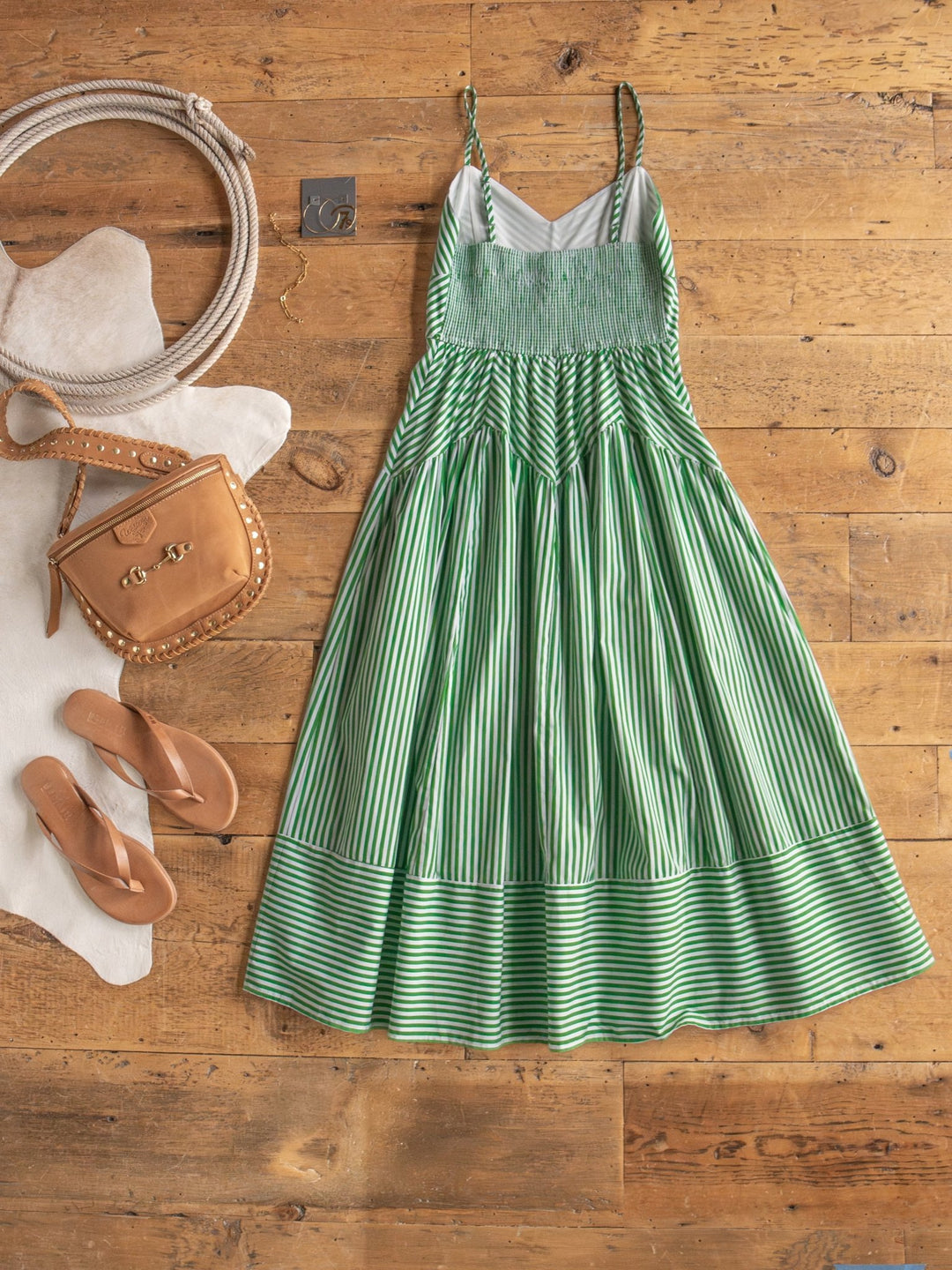 The Lauderhill Green Striped Sleeveless Dress by Savannah Sevens western life{&}style is styled on a wooden floor with tan sandals, a light brown purse, coiled rope, a white hide rug, and gold hoop earrings.