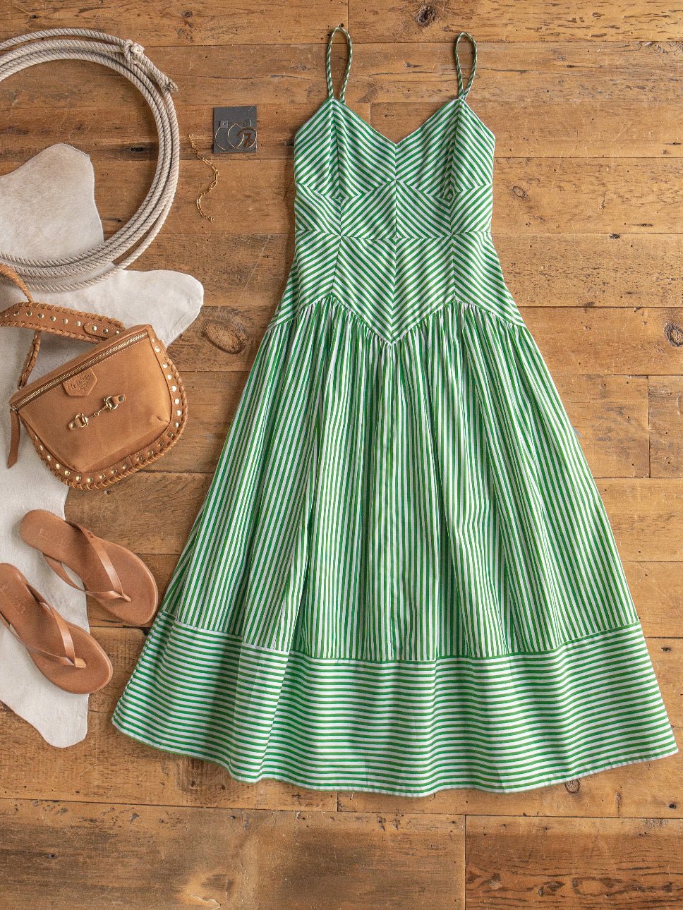 The Lauderhill Green Striped Sleeveless Dress by Savannah Sevens western life{&}style is displayed on a wooden floor alongside tan flip-flops, a tan handbag, coiled rope, a small wallet, and an cowhide rug.