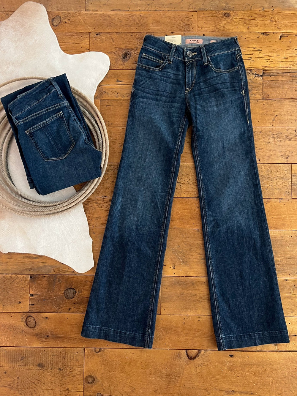 A pair of The Lawson Perfect Rise Trouser Jean by Savannah Sevens western life{&}style is laid flat on a wooden floor, next to a folded Ariat denim trousers and a coiled rope atop a white cowhide rug.