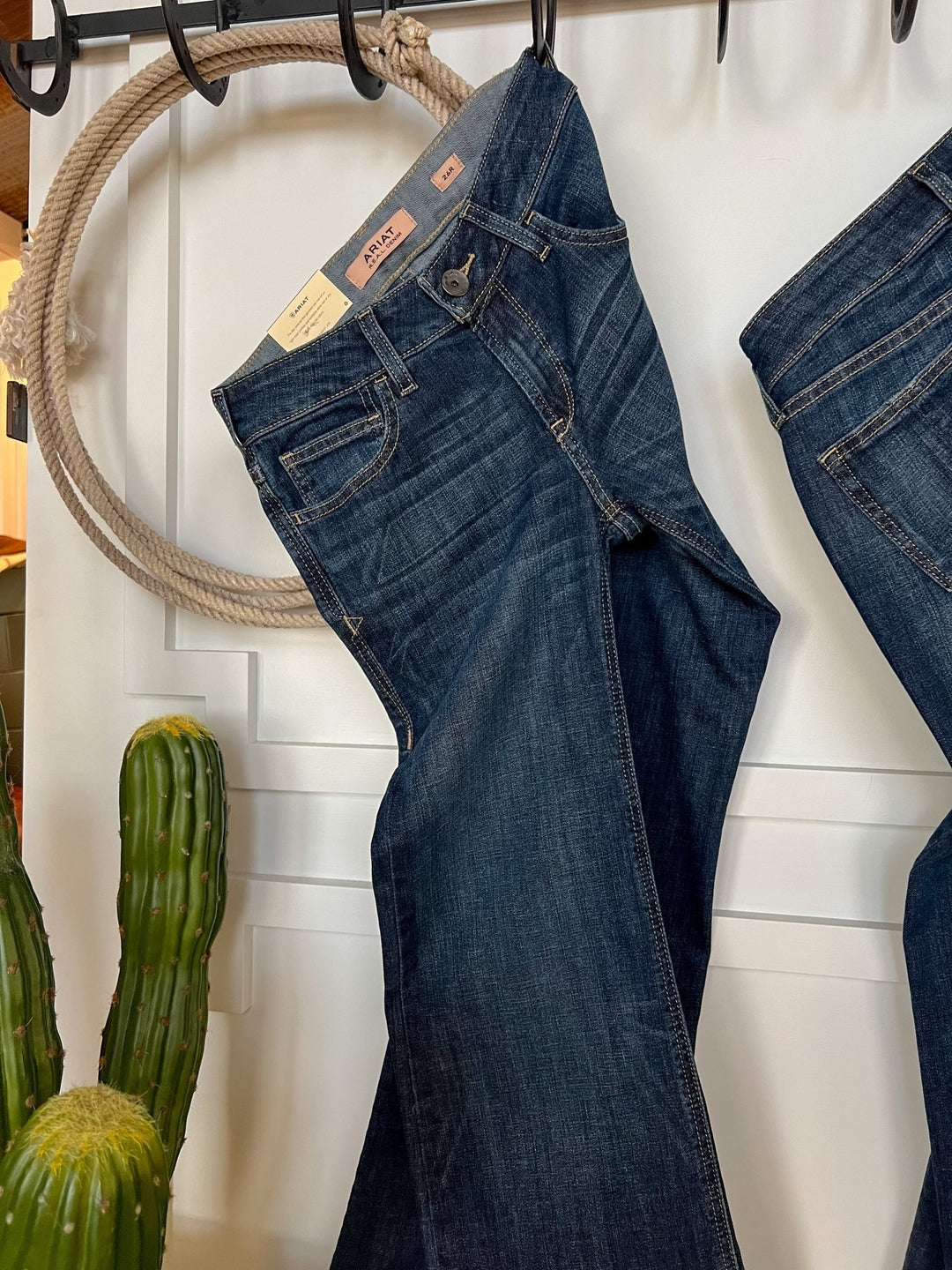 The Lawson Perfect Rise Trouser Jean by Savannah Sevens western life{&}style in a medium dark wash hangs on a black hook against a white wall, with rope and a potted cactus nearby.