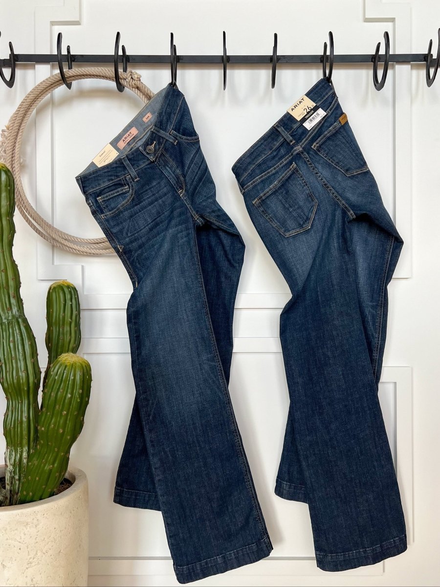 Two pairs of The Lawson Perfect Rise Trouser Jean *Exclusive* by Savannah Sevens western life{&}style hang from black hooks on a white paneled wall, with a potted cactus and decorative rope ring on the left.