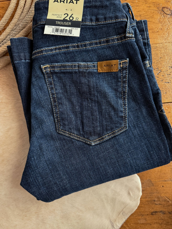 A pair of The Lawson Perfect Rise Trouser Jeans by Savannah Sevens western life{&}style, size 26R, in a medium dark wash with a branded label on the back pocket, folded on a wooden surface and light-colored fabric.
