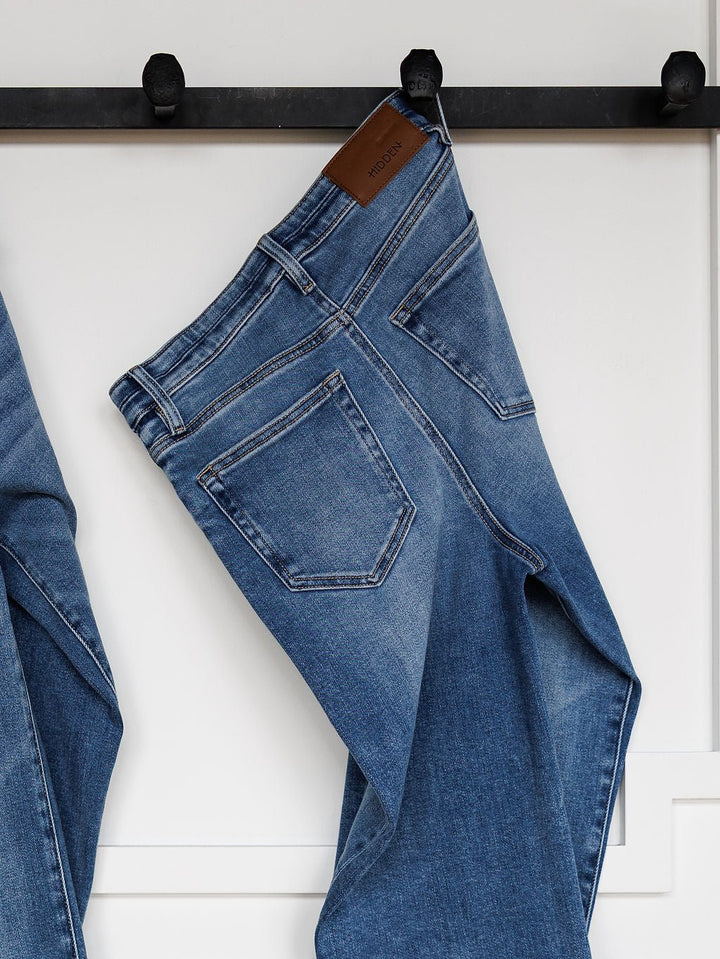 The Layton High Rise Relaxed Straight Leg Jean by Savannah Sevens western life{&}style, featuring visible back pockets and a brown leather brand patch, hangs on a black wall hook against a white background.