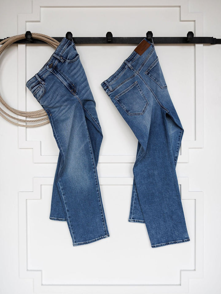 Two pairs of The Layton High Rise Relaxed Straight Leg Jeans by Savannah Sevens western life{&}style hang side by side on a black rod against a white wall with decorative molding; a coiled rope hangs on the left.