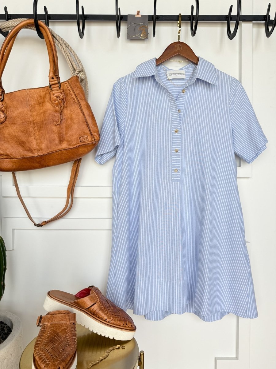 The Leadore Blue Pinstripe Cotton Dress by Savannah Sevens western life{&}style hangs next to a brown leather handbag and woven sandals, all displayed against a white paneled wall.