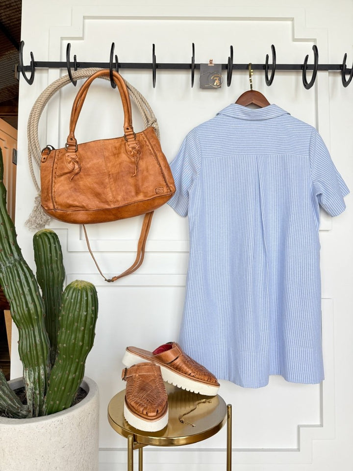 The Leadore Blue Pinstripe Cotton Dress by Savannah Sevens western life{&}style hangs on a black hook rail beside a tan leather handbag, with tan woven platform sandals on a round table next to a tall cactus in a white pot.
