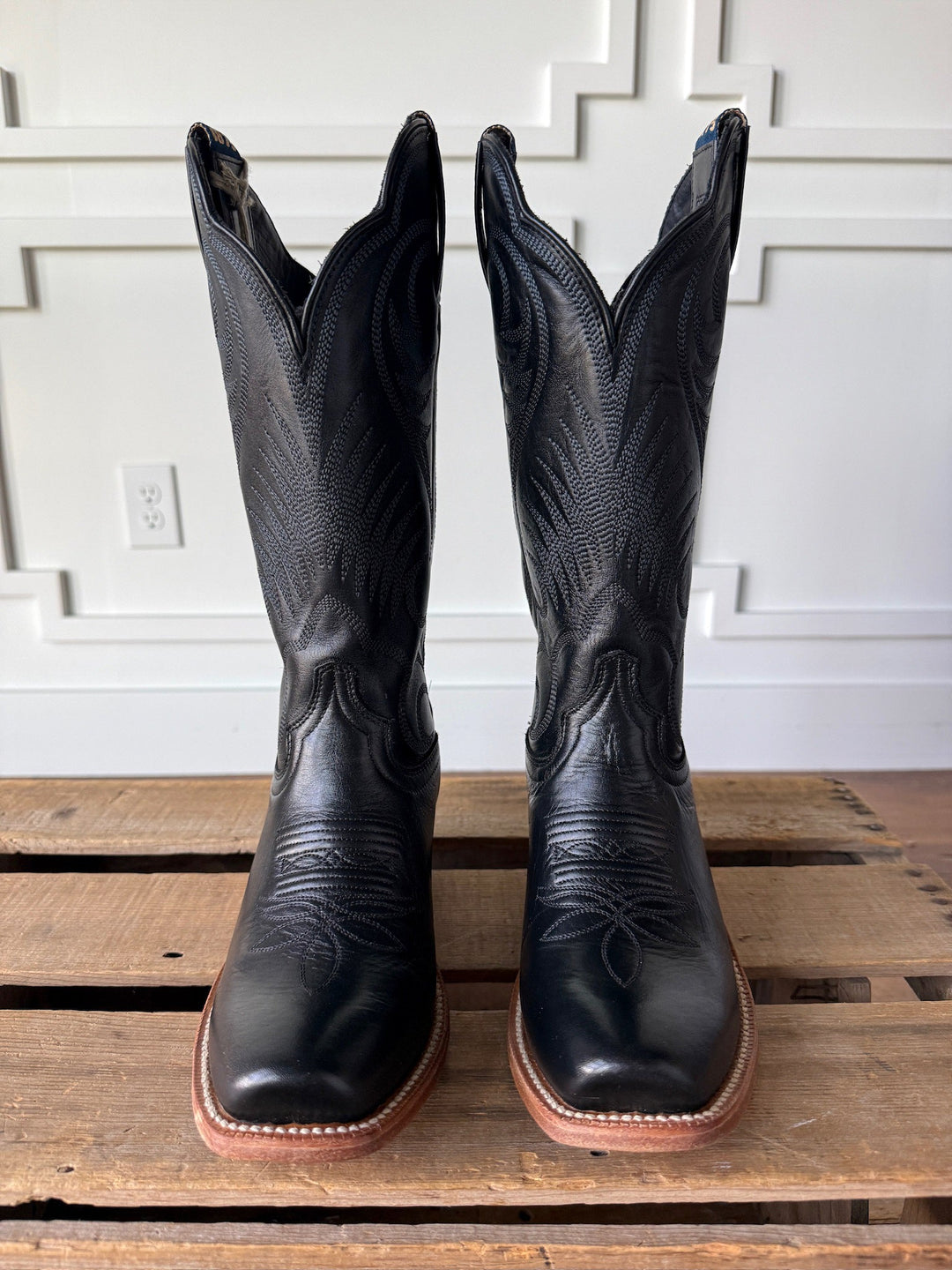 The Leawood Black Western Leather Boots by Savannah Sevens western life{&}style, showcasing intricate stitching and a classic Goodyear welt for added durability, sit side by side on a wooden pallet against a white paneled wall.