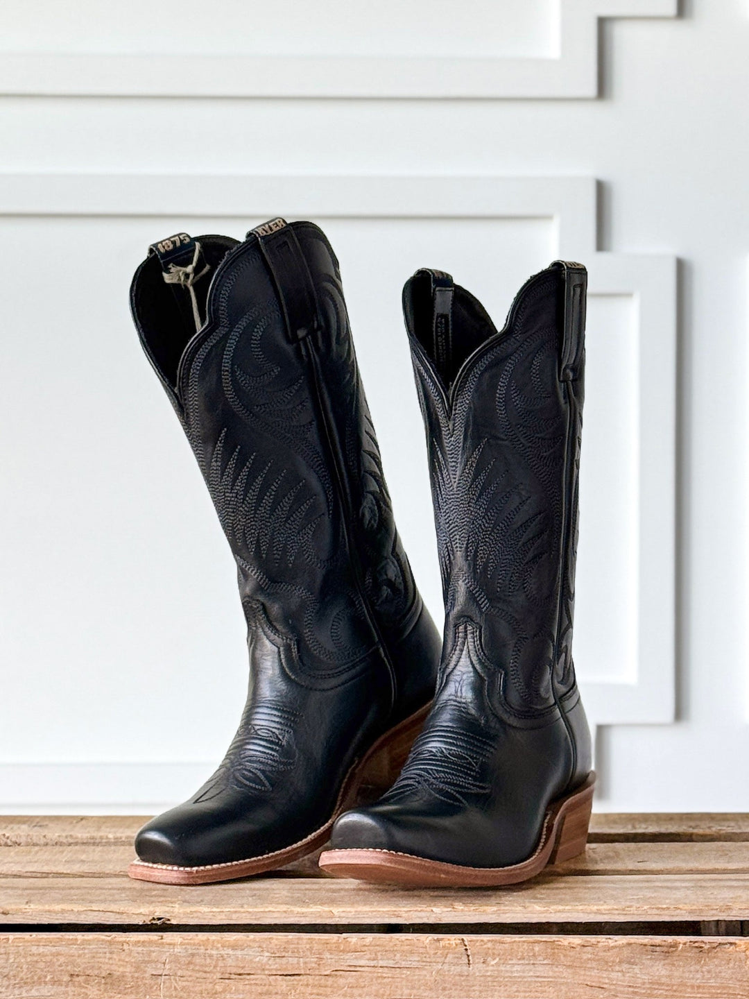 The Leawood Black Western Leather Boot by Savannah Sevens western life{&}style, featuring decorative stitching, sits on a wooden surface in front of a white paneled wall.