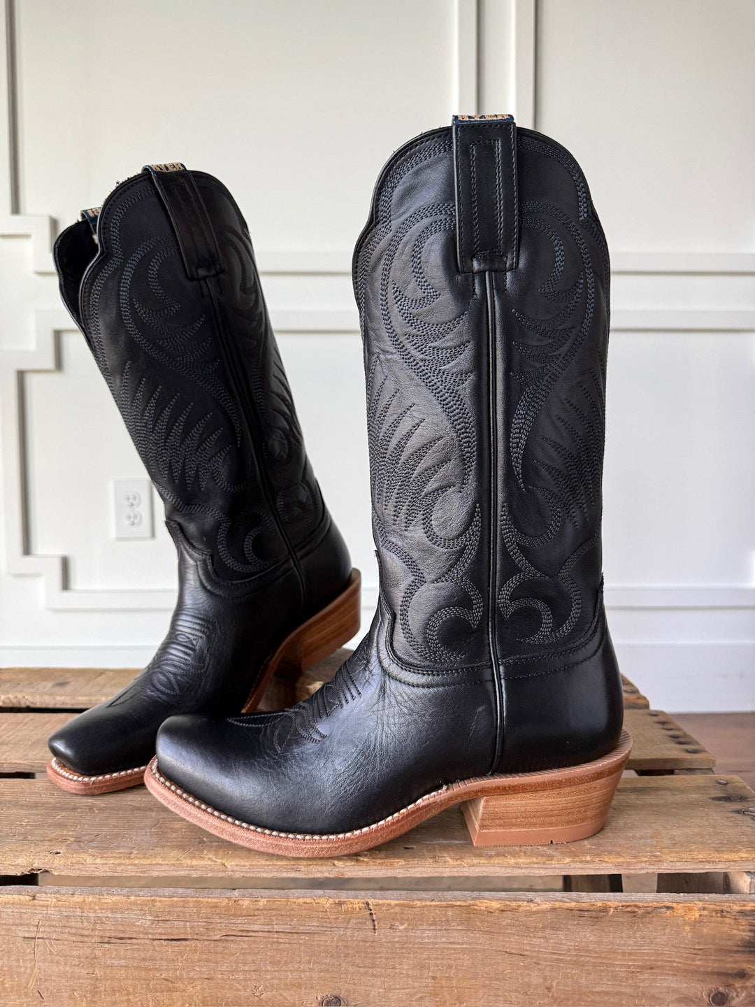 A pair of Leawood Black Western Leather Boots by Savannah Sevens western life{&}style, featuring brown wooden heels, decorative stitching, and classic Goodyear welt construction, displayed on a wooden surface against a white paneled wall.