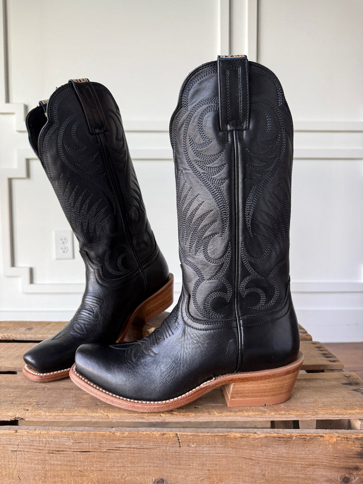 A pair of Leawood Black Western Leather Boots by Savannah Sevens western life{&}style, featuring brown wooden heels, decorative stitching, and classic Goodyear welt construction, displayed on a wooden surface against a white paneled wall.