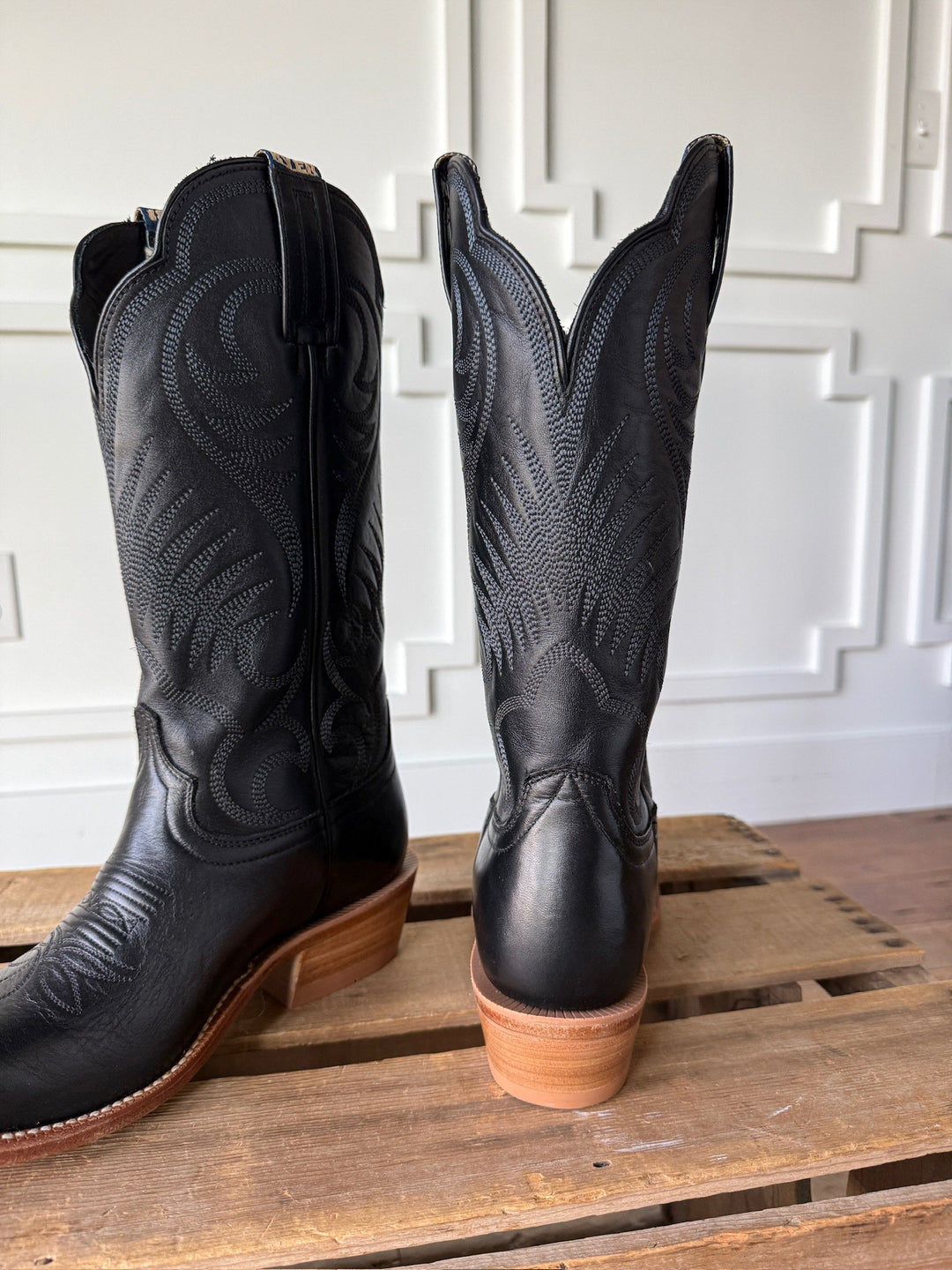 The Leawood Black Western Leather Boot by Savannah Sevens western life{&}style, featuring intricate stitching and a Goodyear welt, is displayed on a wooden surface with a white decorative wall in the background.