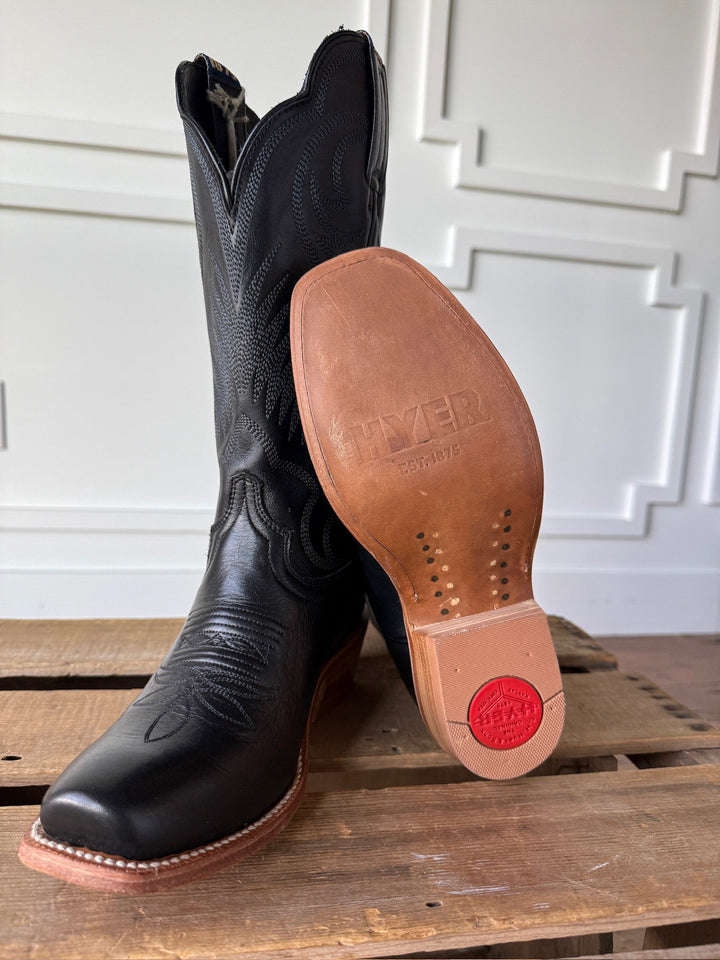 A pair of Leawood Black Western Leather Boots by Savannah Sevens western life{&}style are displayed on a wooden surface; one boot stands upright, while the other leans to showcase its sole with Hyer branding, Goodyear welt construction, and a red circular logo. A white paneled wall serves as the background.