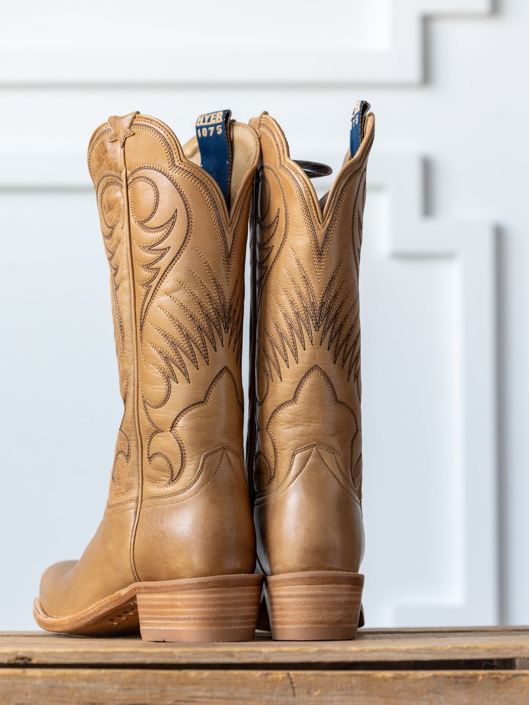 A pair of The Hyer  Leawood Boot {Honey} by Savannah Sevens western life{&}style stands upright on a wooden surface, showcasing detailed stitching and Goodyear leather welt against a white decorative panel background.