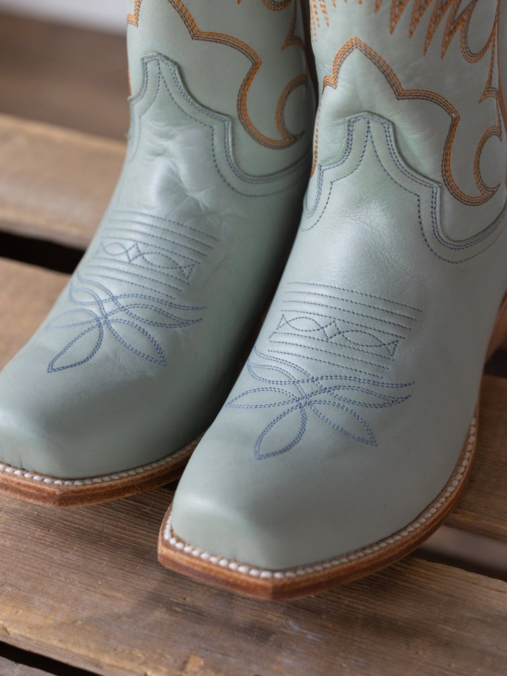 A close-up of The Hyer Leawood Boot {Robin's Egg} by Savannah Sevens western life{&}style showcases light green leather with blue and brown stitching, highlighting the fine craftsmanship of these Western boots on a wooden surface.